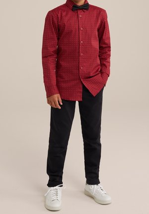 Pojke klädd i en röd skjorta med vita prickar, svart fluga, svarta byxor och vita sneakers som står mot en enfärgad bakgrund.