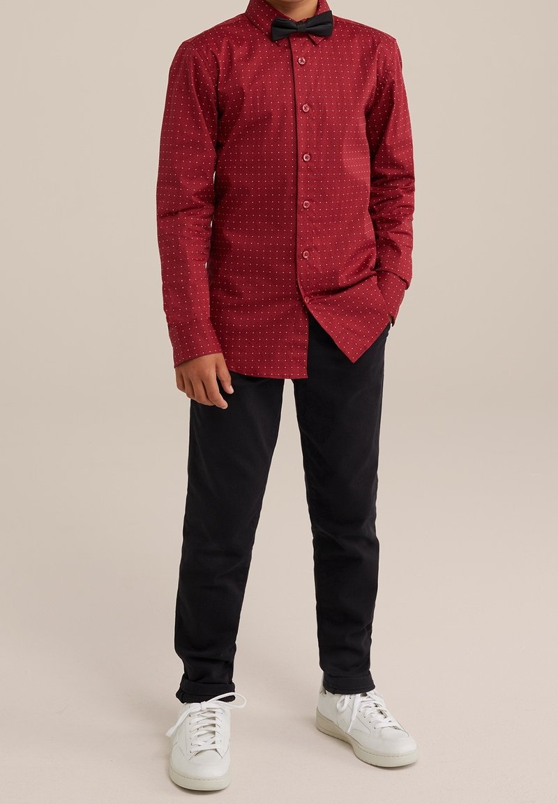 Pojke klädd i en röd skjorta med vita prickar, svart fluga, svarta byxor och vita sneakers som står mot en enfärgad bakgrund.