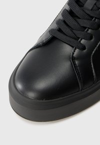 Adidași negri din piele cu vârful rotunjit, talpă din cauciuc plat și șireturi negre. Conțin cusături pe partea superioară pentru un detaliu suplimentar.