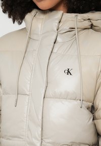 Casaco acolchoado bege claro com acabamento brilhante. Possui gola alta, capuz com cordão ajustável e logótipo frontal em preto.