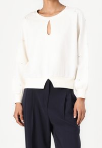 Vrouw die een crèmekleurige top met lange mouwen en sleutelgat-halslijn draagt, gecombineerd met zwarte, brede, geplooide pantalon, staand tegen een witte achtergrond.