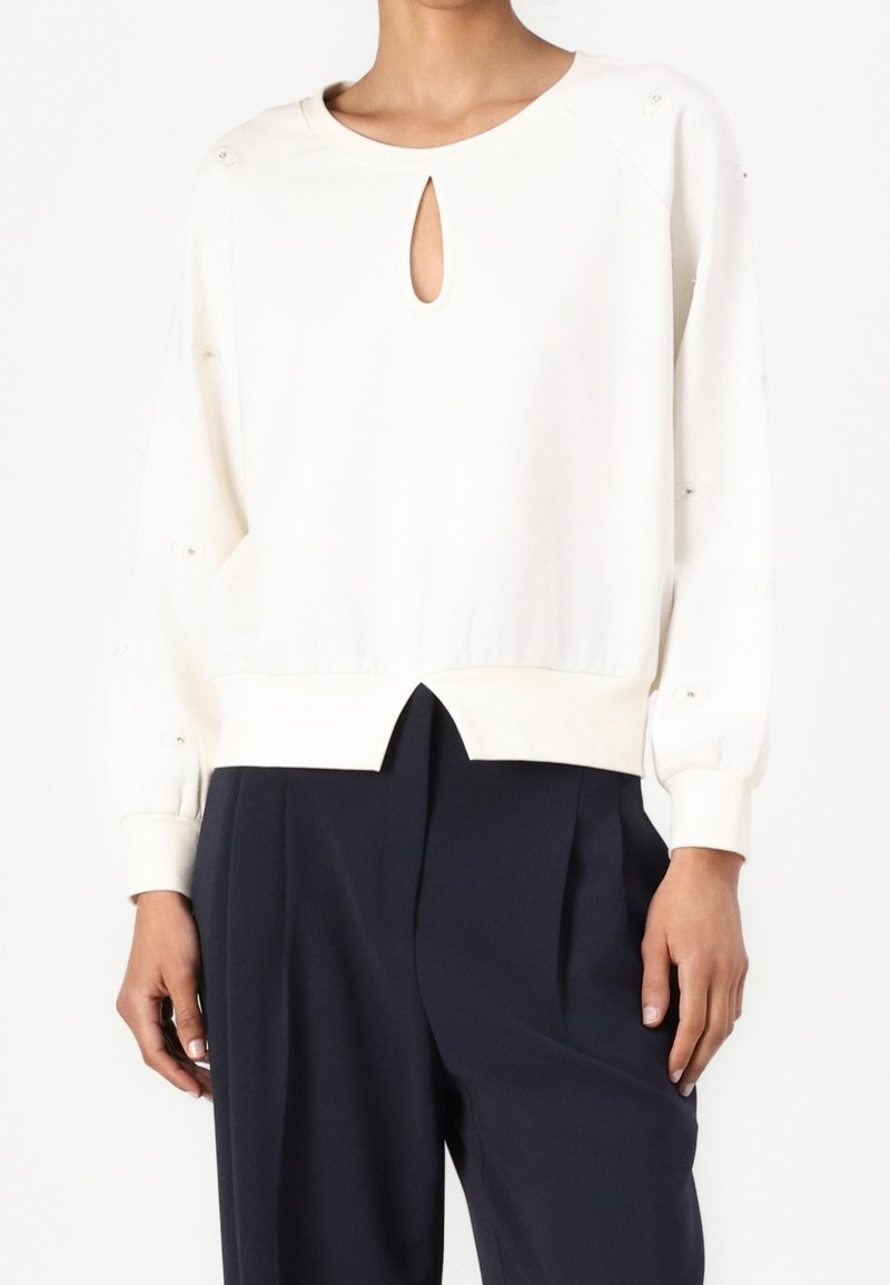 Vrouw die een crèmekleurige top met lange mouwen en sleutelgat-halslijn draagt, gecombineerd met zwarte, brede, geplooide pantalon, staand tegen een witte achtergrond.