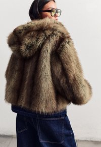 Bruine nepbontjas met een capuchon, voorzien van een boxy ontwerp en een gestructureerde afwerking. Gecombineerd met donkere denim broeken.