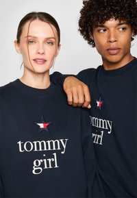 Mørkeblå sweatshirts med rund hals, der har hvid broderet "tommy girl" tekst og et stjernesymbol med røde, hvide og blå detaljer.
