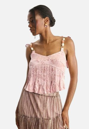 Mujer con cabello trenzado lleva una blusa rosa con flores y pliegues, con tirantes decorativos y una falda marrón texturizada, mirando hacia un lado.