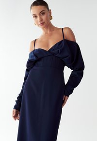 Námořnické šaty s odhalenými rameny, s přiléhavým korzetem a objemovými rukávy. Vyrobené z hladkého materiálu, s drapérií kolem výstřihu.