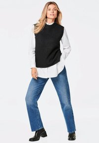 Schwarzer gestrickter ärmelloser Pullover über einem weißen Kragenhemd, kombiniert mit blauen geraden Jeans und schwarzen Absatzstiefeln.