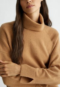 Maglione beige lavorato a maglia con un alto colletto a coste. Presenta maniche testurizzate e costine orizzontali sulle spalle. Materiale morbido e accogliente.