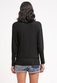 Femme aux cheveux ondulés châtains mi-longs, portant un haut noir à manches longues et un short en jean retroussé, vue de dos sur un fond blanc.