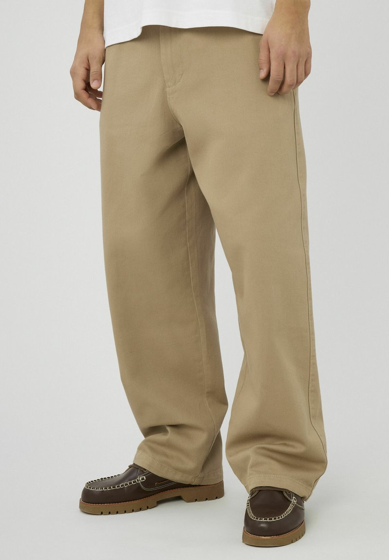 Pantalon beige à jambes larges en tissu de coton épais, avec une surface texturée, assorti à des mocassins marron foncé avec des accents de couture.