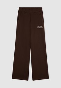 Brązowe szerokie spodnie dresowe z elastyczną talią, bocznymi kieszeniami oraz białym logo "Stockholm Leisure Club" na lewej udzie.