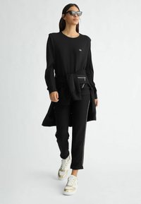 Maglietta nera a maniche lunghe con scollo rotondo, abbinata a pantaloni neri con striscia laterale e sneaker dorate con suole bianche.