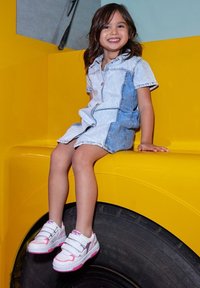 Baskets blanches et roses avec des bandes Velcro et des accents argentés ; robe chemise en denim ; assise sur une surface jaune près d'un pneu noir.