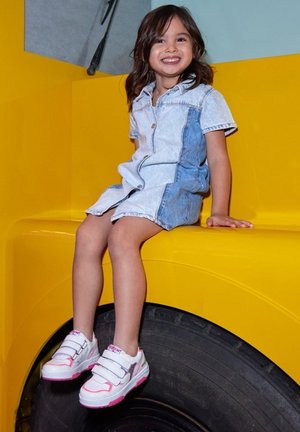 White and pink sneakers with velcro straps and silver accents; denim shirt dress; seated on a yellow surface near a black tire.