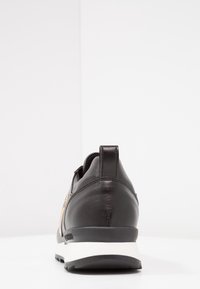 Black leather sneaker with a light tan accent, rounded shape, textured finish, and white midsole. Features a pull tab at the heel.