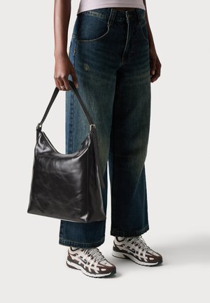Black leather handbag with a curved shape, a flat bottom, and a long adjustable strap, held by a person in loose blue jeans and sneakers.