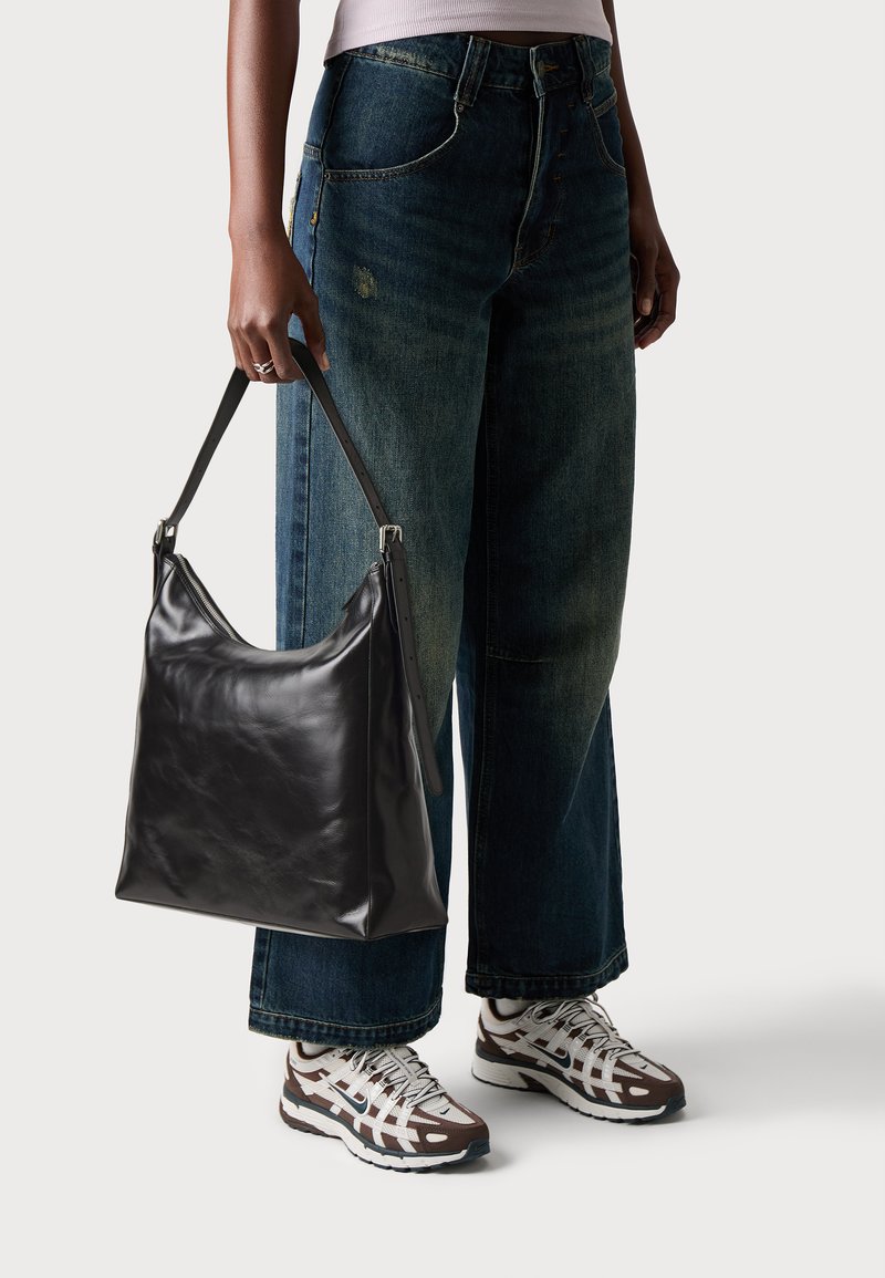 Sac en cuir noir de forme courbée, avec un fond plat et une longue bandoulière ajustable, tenu par une personne en jean bleu décontracté et en baskets.