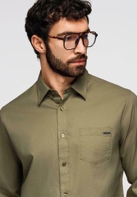 Homme avec barbe et cheveux bouclés portant des lunettes écaille de tortue et une chemise boutonnée vert olive avec une poche poitrine, regardant vers le bas à droite.