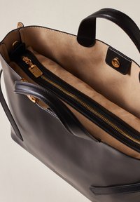 Schwarze Lederhandtasche mit glatter Oberfläche und goldenen Beschlägen. Das Innere verfügt über ein hellbraunes Wildlederfutter und ein Reißverschlussfach zur besseren Organisation.