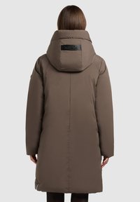 Pikk pruun polsterdatud mantel suure kapuutsiga ja musta brändi sildiga. Sile tekstuur, sirge lõige ning küljetaskud.