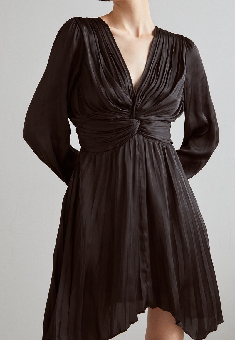 Femme portant une robe plissée noire à manches longues, un col en V profond et un détail de nœud torsadé à la taille.
