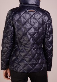 Mörkblå quiltad puffjacka med hög krage, med en slät, glänsande yta och diagonalt sömnadsmönster. Bruna accenter i metall vid nacken.