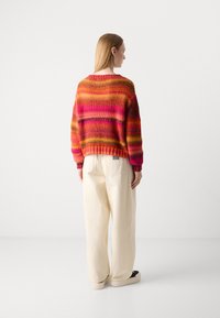 Multikleurige gebreide trui met horizontale strepen in roze, oranje en geel. Gecombineerd met crèmekleurige wijde broek en donkere schoenen.