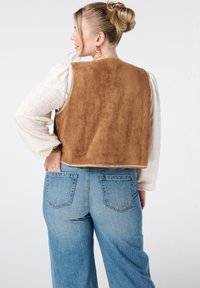Bruine faux-suede crop vest met een gladde textuur, gecombineerd met een witte broderie anglaise blouse en high-waisted blauwe spijkerbroeken.