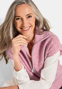 Gilet zippé rose avec texture côtelée, superposé sur une blouse blanche. Il présente un col montant et des fermetures éclair en argent.