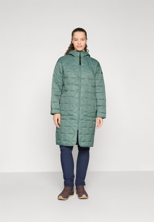 Manteau long en vert matelassé avec capuche, doté d'une fermeture zippée à l'avant, d'un motif triangulaire et de poches latérales, porté par-dessus un pantalon bleu marine et des bottes marron.
