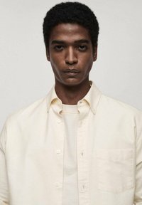 Lichtbeige knoopsluiting overhemd met een kraag en borstzak, gedragen over een lichtbruin t-shirt. Glad stof, minimalistisch ontwerp.