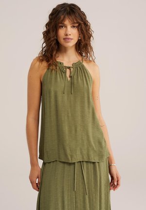 Kobieta o kręconych włosach, ubrana w oliwkowozielony top bez rękawów z dekoltem halter i dopasowaną spódnicę na jednolitym tle.