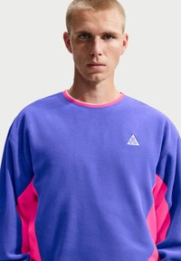 Νεαρός με κοντά ξανθά μαλλιά που φοράει φούτερ σε μωβ και ροζ αποχρώσεις με λευκό λογότυπο ACG στο στήθος, μπροστά σε ένα απλό ανοιχτόχρωμο φόντο.