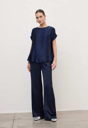 Vrouw draagt een loszittend marineblauw zijden topje en bijpassende wijde broek, staand tegen een effen lichte muur en vloer.