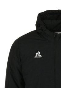 Veste zippée noire avec un col haut, ornée d'un logo blanc sur la poitrine gauche. Fabriquée dans un matériau lisse et léger.