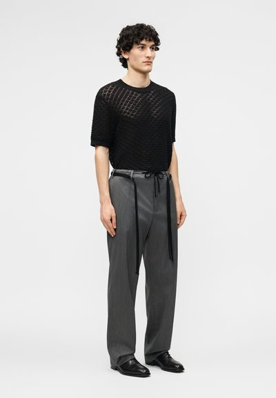 Νέος άνδρας που φοράει μαύρο πουλόβερ με κοντά μανίκια από διχτυωτό ύφασμα, φαρδιά γκρι παντελόνια με μαύρα μακριά κορδόνια και μαύρα επίσημα παπούτσια, στέκεται μπροστά σε λευκό φόντο.