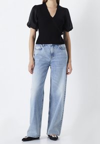 Haut portefeuille noir côtelé avec manches bouffantes, associé à un jean bleu clair à jambes larges. Design simple, texture moyenne et contraste de couleurs uniforme.