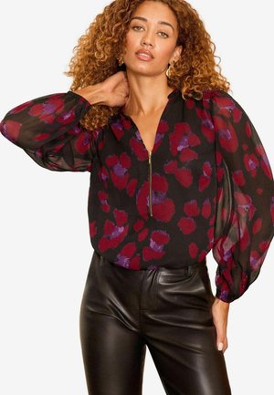 Schwarze Bluse mit mehrfarbigem Blumenmuster in Rot und Lila, ausgestattet mit einem Reißverschluss-Halsausschnitt und transparenten Puffärmeln, kombiniert mit schwarzen Lederhosen.