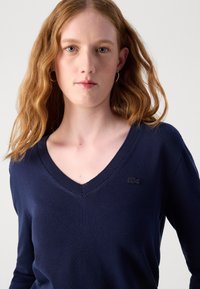 Maglione blu navy a collo a V realizzato in materiale morbido, con un ricamo logo discreto, scollatura a costine e una forma aderente.
