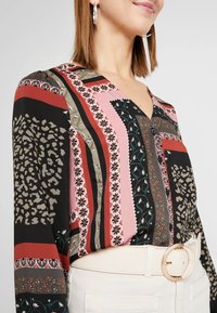 Donna che indossa una blusa a stampa floreale e animale multicolore con bottoni neri, abbinata a pantaloni bianchi e una cintura beige con fibbia rotonda.