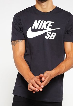 Mand iført en sort Nike SB t-shirt med hvidt logo, hænderne foldet foran, og en delvis tatovering synlig på hans øverste venstre arm.