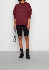 Sudadera oversized color marrón burdeos con un bolsillo frontal, combinada con pantalones cortos de ciclismo negros y botas de cordones negras. El accesorio incluye un bolso estampado.
