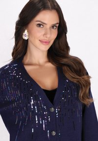 Marineblauwe cardigan met sequins in verschillende kleuren, een diepe V-halslijn en zilverkleurige knopen aan de voorkant. Textuurgebreid ontwerp.