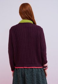 Frau mit langem roten Haar trägt einen dunkelvioletten gestrickten Pullover mit limonengrünem Kragen und pinkem Rand, kombiniert mit einem grünen karierten Rock, Rückansicht.