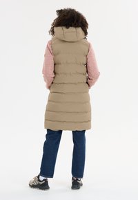 Beige puffer vest dengan penutup kepala, menampilkan desain quilted. Lengan panjang berwarna pink dan jeans biru tua, dilengkapi dengan sepatu beige dan hitam.