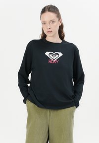 Black long-sleeve t-shirt with a white heart logo and "ROXY" in pink. Features a round neck and relaxed fit, paired with green corduroy pants.