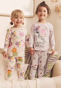 Deux enfants se tiennent la main, vêtus de pyjamas. L'un porte un haut rose clair orné d'une licorne et de motifs pastel, l'autre porte des motifs floraux.