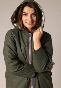 Veste verte olive avec une capuche doublée, des accents métalliques et une fermeture éclair rouge, blanche et bleue contrastante. Texture lisse, coupe décontractée.