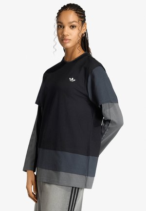 Joven con trenza lleva una camisa Adidas de capas negra, azul oscuro y gris y pantalones grises con rayas negras, de pie sobre fondo blanco.