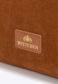 Bruine suède tas met een goud-gegraveerd WITTCHEN-logo op een leren label, met een gladde textuur en rechthoekige vorm.